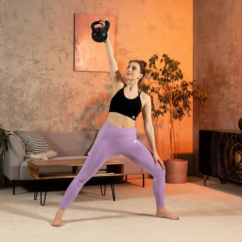 Kobieta trenująca w domu, prezentuje technikę ćwiczenia kettlebell windmill – z ciężarem uniesionym nad głową i szerokim rozstawem nóg.