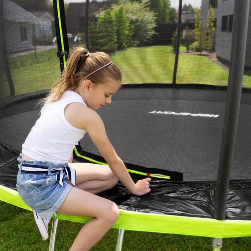 Dziewczynka w białej koszulce i jeansowych szortach zapina zamek siatki ochronnej w trampolinie ogrodowej – bezpieczna trampolina ogrodowa czy warto dla dzieci.