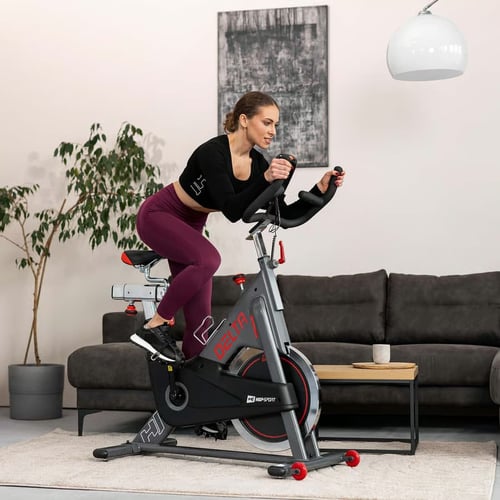 Kobieta wykonująca intensywny trening na rowerze spinningowym Hop-Sport Delta w salonie – dynamiczna pozycja jazdy idealnie ilustruje różnicę w wyborze: rower treningowy czy spinningowy
