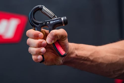 Mężczyzna ćwiczy nadgarstek za pomocą regulowanego ściskacza do rąk z metalową sprężyną marki Hop-Sport; realistyczna prezentacja sprzętu i możliwych efektów treningu siły chwytu.